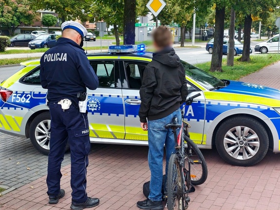 Przy radiowozie stoi policjant i młody chłopak z rowerem.