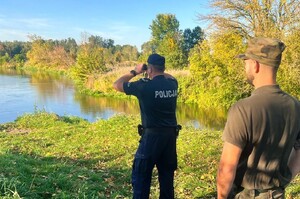 Nad wodą, stoi policjant i patrzy w stronę zarośli przez lornetkę. Za nim stoi strażnik rybacki.