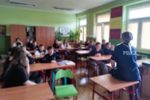Klasa w szkole. Uczniowie siedzą w ławkach, przed nimi stoi policjantka, mówi do nich.