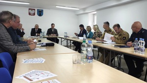 W pomieszczeniu są dwa długie stoły, przy których siedzą uczestnicy spotkania. W szczycie siedzi starosta i jego zastępca.