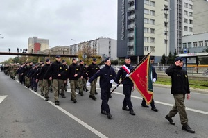 Uczniowie w mundurach idą w defiladzie z pocztem sztandarowym wzdłuż ulicy.