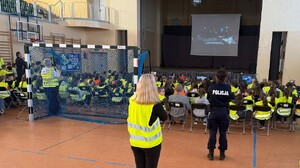 Sala gimnastyczna, na krzesełkach ustawionych w rzędach siedzą uczniowie w kamizelkach odblaskowych. Patrzą na ekran przed sobą, na którym wyświetlany jest film profilaktyczny. Z tyłu za uczniami stoi policjantka.